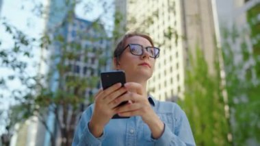Gözlüklü beyaz kadın şehrin caddesinde dikiliyor ve akıllı telefon kullanıyor. Arka planda gökdelenler var. İletişim, iş günü, yoğun yaşam konsepti. Yavaş çekim