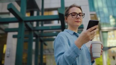 Gözlüklü beyaz kadın sokakta duruyor, akıllı telefon kullanıyor ve kahve içiyor. Yörünge atışı. Arka planda gökdelenler var. İletişim, iş günü, yoğun yaşam konsepti.