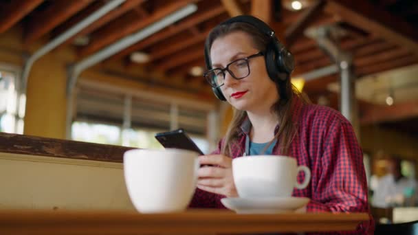 带着耳机的女人正在咖啡店里听着音乐 听着音乐 听着音乐 边听边喝咖啡 在咖啡店或餐馆欣赏播客 声音或歌曲的女性或自由职业者 — 图库视频影像
