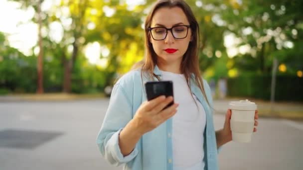 スマートフォンとコーヒーを持った幸せな女性は 夏の夕方に街頭に立っています オービットショット — ストック動画