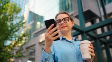 Gözlüklü beyaz kadın sokakta duruyor, akıllı telefon kullanıyor ve kahve içiyor. Yörünge atışı. Arka planda gökdelenler var. İletişim, iş günü, yoğun yaşam konsepti.