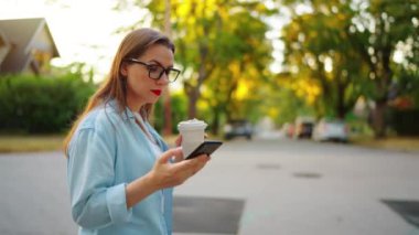 Akıllı telefonlu ve elinde kahve olan mutlu bir kadın yaz akşamı mahallede dolanıyor. Yavaş çekim