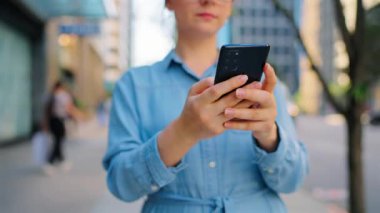 Gözlüklü beyaz kadın şehirde dolanıyor ve akıllı telefon kullanıyor. Arka planda gökdelenler var. İletişim, iş günü, yoğun yaşam konsepti. Yavaş çekim