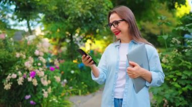 Dizüstü bilgisayarı ve akıllı telefonu olan mutlu bir kadın çiçek açan bahçede ya da işten sonra yeşil sokakta duruyor..