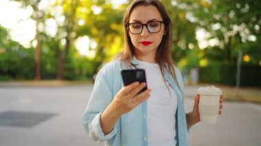 Akıllı telefonu ve kahvesi olan mutlu kadın yaz akşamları sokakta ayakta duruyor. Yörünge çekimi, yavaş çekim.