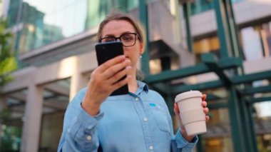 Gözlüklü beyaz kadın sokakta duruyor, akıllı telefon kullanıyor ve kahve içiyor. Yörünge atışı. Arka planda gökdelenler var. İletişim, iş günü, yoğun yaşam konsepti. Yavaş çekim