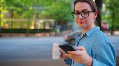 Beyaz gözlüklü kadın cadde boyunca yürüyor, akıllı telefon kullanıyor ve kahve içiyor. Arka planda gökdelenler var. İletişim, iş günü, yoğun yaşam konsepti.