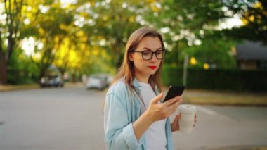 Akıllı telefonu ve kahvesi olan mutlu kadın yaz akşamları sokakta ayakta duruyor. Yörünge atışı
