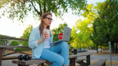 Kadın parkta oturuyor, bir dizüstü bilgisayar üzerinde çalışıyor ve kahve içiyor. Yavaş çekim