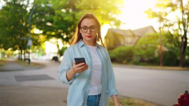Güneşli bir yaz gününde yeşil caddede yürürken akıllı telefon kullanan mutlu bir kadının portresi. Yavaş çekim