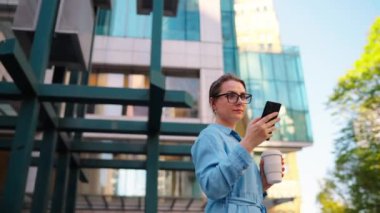 Gözlüklü beyaz kadın sokakta duruyor, akıllı telefon kullanıyor ve kahve içiyor. Yörünge atışı. Arka planda gökdelenler var. İletişim, iş günü, yoğun yaşam konsepti. Yavaş çekim