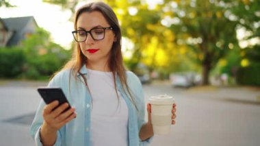 Akıllı telefonu ve kahvesi olan mutlu kadın yaz akşamları sokakta ayakta duruyor. Yörünge atışı. Yavaş çekim