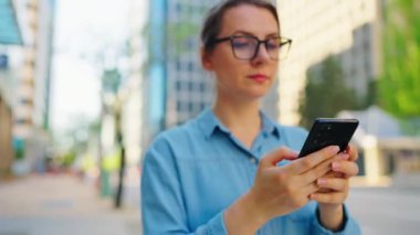 Gözlüklü beyaz kadın şehirde dolanıyor ve akıllı telefon kullanıyor. Arka planda gökdelenler var. İletişim, iş günü, yoğun yaşam konsepti.