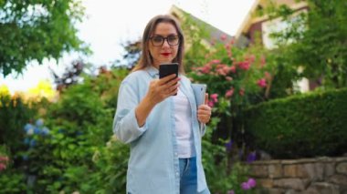 Dizüstü bilgisayarı ve elinde akıllı telefonu olan mutlu bir kadın çiçek açan bahçede ya da işten sonra Green Street 'te duruyor. Yörünge atışı