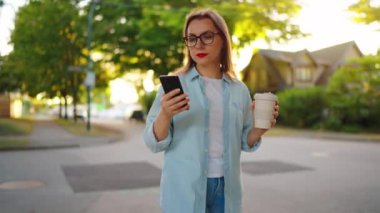 Akıllı telefonlu ve elinde kahve olan mutlu bir kadın yaz akşamı mahallede dolaşıyor..