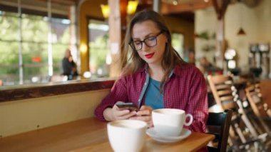 Akıllı telefon kullanan ve kafede kahve içen bir kadın. Kentsel yaşam tarzı, sosyal medyada mesajlaşmak, mobil teknolojiden hoşlanmak, kahve dükkanında dinlenmek. Yavaş çekim