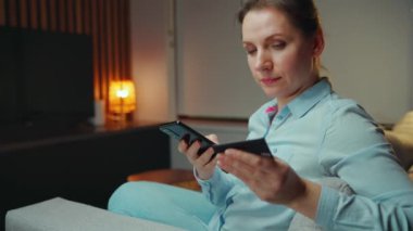 Bir kadın evde dinlenirken online alışveriş için akıllı telefonunu ve kredi kartını kullanırken gülümsüyor.