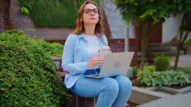 Kadın parkta dizüstü bilgisayar ve kahveyle oturuyor ve yaz boyunca çalışıyor..