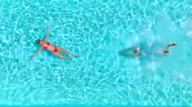 Çift havuzda rahatlar. Kadın sırt üstü yatarken bir erkek havuza dalıp yüzerken yukarıdan aşağı bakıyor. Yavaş çekim