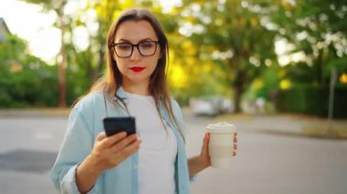 Akıllı telefonu ve kahvesi olan mutlu kadın yaz akşamları sokakta ayakta duruyor. Yörünge atışı.