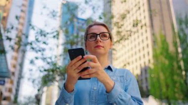 Gözlüklü beyaz kadın sokakta dikiliyor ve akıllı telefon kullanıyor. Arka planda gökdelenler var. İletişim, iş günü, yoğun yaşam konsepti. Yörünge atışı