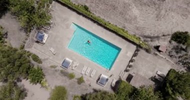 Kırmızı mayo giymiş bir kadının havuzda sırtüstü yüzüşü. Rahatlatıcı konsept, yaz yaşam tarzı. Havuz ve çöl manzarası. Kamera indirir ve döndürür