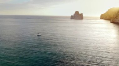 Yelkenlinin kayalık kıyıya yakın derin mavi bir denizde yelken açtığını gösteren hava görüntüleri. Drone 'dan deniz burnu. İtalya, Sardinya 'da gün batımı. Seyahat videosu