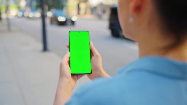 Kadın cadde boyunca yürüyor ve yeşil ekranlı akıllı telefon kullanıyor. İletişim, iş günü, yoğun yaşam konsepti.