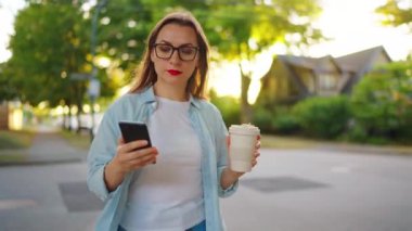 Akıllı telefonlu ve elinde kahve olan mutlu bir kadın yaz akşamı mahallede dolaşıyor..