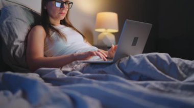 Gözlüklü kadın geceleri yatakta yatarken dizüstü bilgisayarda çalışıyor. İnternet bağımlısı veya uykusuzluk konsepti. 
