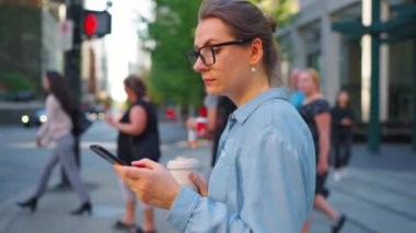 Beyaz gözlüklü kadın caddenin karşısına geçiyor, akıllı telefon kullanıyor ve kahve içiyor. Arka planda gökdelenler var. İletişim, iş günü, yoğun yaşam konsepti. Yavaş çekim