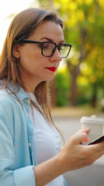 Elinde akıllı telefon ve kahveyle mutlu bir kadın yaz akşamı mahallede dolaşıyor. Ağır çekim. Dikey video