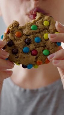 Aç bir kadın renkli cipsli kurabiyeleri yakın plan ısırıp çiğniyor. Kadın sağlıksız yemek konsepti. Yiyecek kalorisi. Dikey video