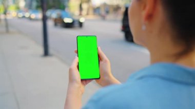 Kadın cadde boyunca yürüyor ve yeşil ekranlı akıllı telefon kullanıyor. İletişim, iş günü, yoğun yaşam konsepti. Yavaş çekim