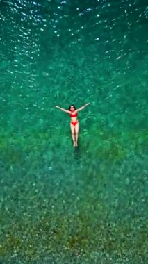 Kırmızı mayo giymiş bir kadının havadan görünüşü kristal berrak deniz sularında rahatlığı somutlaştırarak rahatça süzülür. Yaz tatilleri. Dikey video