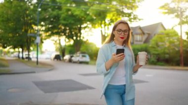 Akıllı telefonlu ve elinde kahve olan mutlu bir kadın yaz akşamı mahallede dolaşıyor..