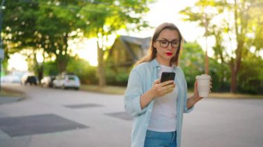 Elinde akıllı telefon ve kahveyle mutlu bir kadın yaz akşamı mahallede dolaşıyor. Yavaş çekim