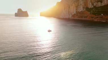 Yelkenlinin kayalık kıyıya yakın derin mavi bir denizde yelken açtığını gösteren hava görüntüleri. Drone 'dan deniz burnu. İtalya, Sardinya 'da gün batımı. Seyahat videosu