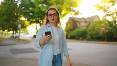 Güneşli bir yaz gününde yeşil caddede yürürken akıllı telefon kullanan mutlu kadının portresi..