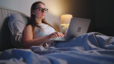 Gözlüklü kadın geceleri yatakta yatarken dizüstü bilgisayarda çalışıyor. İnternet bağımlısı veya uykusuzluk konsepti. 