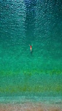 Kırmızı mayo giymiş bir kadının havadan görünüşü berrak deniz sularında sırtüstü yüzüyor, rahatlatıcı bir şekilde. Yaz tatilleri. Dikey video