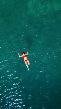 Kırmızı mayo giymiş bir kadının havadan görünüşü berrak deniz sularında sırtüstü yüzüyor, rahatlatıcı bir şekilde. Yaz tatilleri. Dikey video