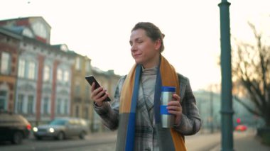 Beyaz ceketli bir iş kadını, sabahın erken saatlerinde şehirde dolaşıyor, kahve içiyor ve akıllı telefon kullanıyor, ağır çekimde. İletişim, iş günü, yoğun yaşam konsepti.