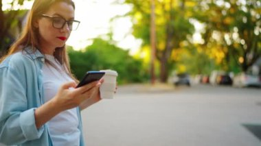 Elinde akıllı telefon ve kahveyle mutlu bir kadın yaz akşamı mahallede dolaşıyor. Yavaş çekim