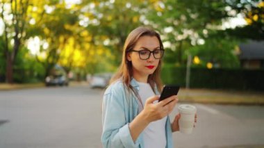 Akıllı telefonu ve kahvesi olan mutlu kadın yaz akşamları sokakta ayakta duruyor. Yörünge atışı. Yavaş çekim