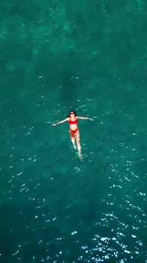 Kırmızı mayo giymiş bir kadının havadan görünüşü, kristal berrak deniz sularında rahatlığı somutlaştırmak için zahmetsizce yüzüyor. Yaz tatilleri. Dikey video