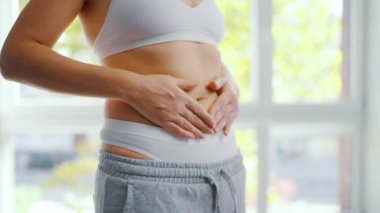 Kadın pantolonunu giyer ve karnını okşar. Sağlıklı sindirim ve bağırsak fonksiyonu kavramı. Kişinin vücut pozitifliğini göstermesi, kendini kabullenme ve refahı vurguluyor.