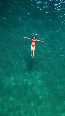 Kırmızı mayo giymiş bir kadının havadan görünüşü, kristal berrak deniz sularında rahatlığı somutlaştırmak için zahmetsizce yüzüyor. Yaz tatilleri. Dikey video