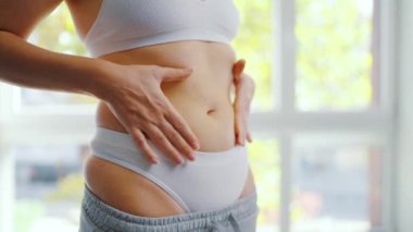 Kadın pantolonunu giyer ve karnını okşar. Sağlıklı sindirim ve bağırsak fonksiyonu kavramı. Kişinin vücut pozitifliğini göstermesi, kendini kabullenme ve refahı vurguluyor.