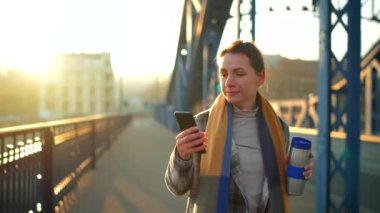 Beyaz ceketli bir iş kadını, sabahın erken saatlerinde şehirde dolaşıyor, kahve içiyor ve akıllı telefon kullanıyor. İletişim, iş günü, yoğun yaşam konsepti.